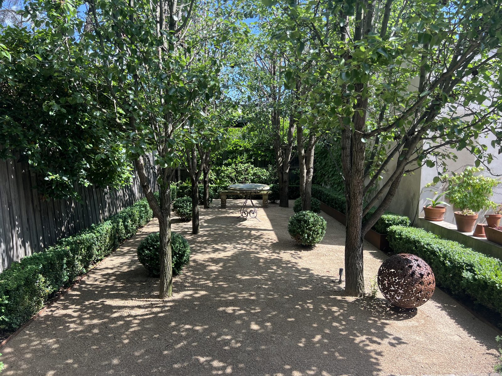 A gravel courtyard walk beneath pleached pears, box hedges and a corten sphere sculpture